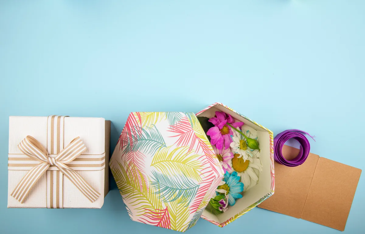 top-view-gift-box-tied-with-bow-gift-box-filled-with-colorful-chrysanthemum-flowers-with-daisy-purple-ribbon-with-small-postcard-blue-background.webp